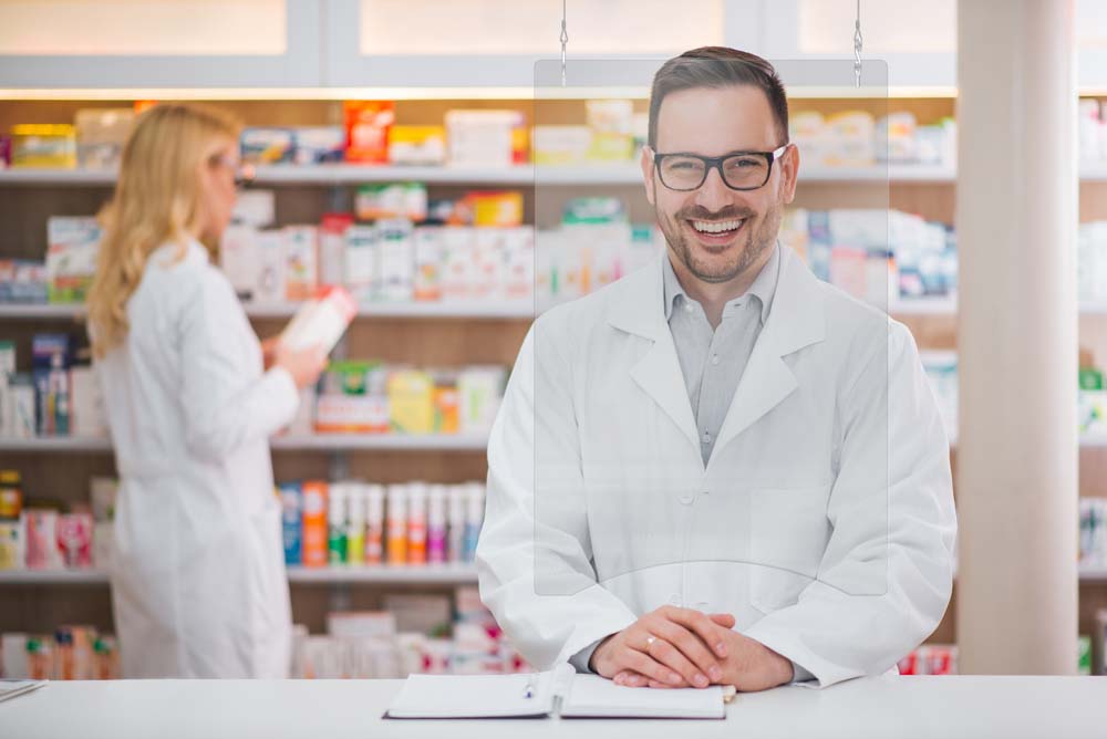 Portrait of a handsome pharmacist at the counter of a drugstore, female colleague working in the background.